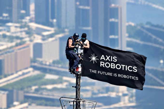 Two people in tactical gear sit atop a pole holding an 'AXIS ROBOTICS - THE FUTURE IS ROBOTICS' flag, with a hazy cityscape and bridge visible in the background.