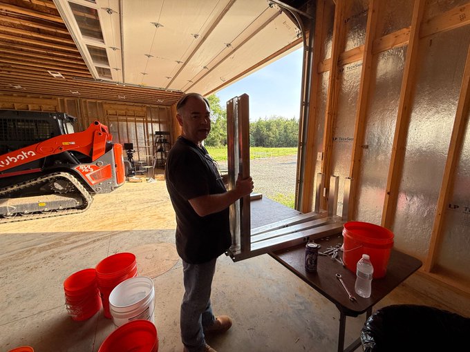 A man wearing safety glasses stands inside an unfinished garage holding a window frame, with a Kubota skid-steer loader, red and white buckets, and tools visible in the workspace.