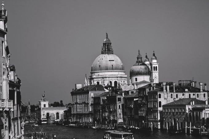 A black-and-white photograph of Venice, Italy, showing the Grand Canal with boats and the prominent dome of the Basilica di Santa Maria della Salute in the background.