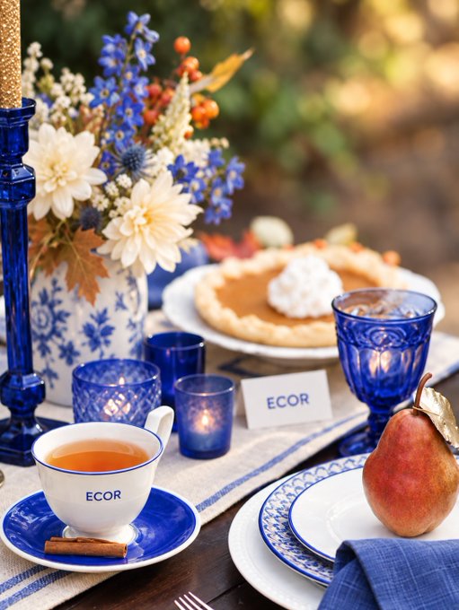 An autumn-themed table setting featuring blue and white china branded 'ECOR', blue glassware, a vase of blue and white flowers, pumpkin pie with whipped cream, and a pear.