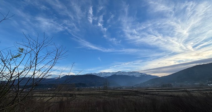 A scenic landscape photograph showing snow-capped mountains in the distance under a bright blue sky with wispy cirrus clouds, with bare tree branches and dry grass in the foreground.
