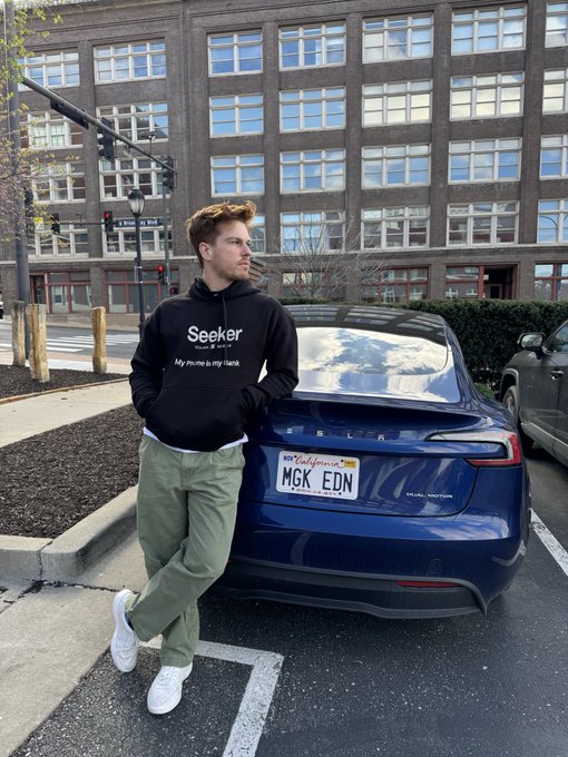 A young man leans against a blue Tesla with a California license plate reading 'MGK EDN'; he wears a black hoodie with 'Seeker Group' and 'My Home is my Tank' printed on it, along with green pants and white sneakers.