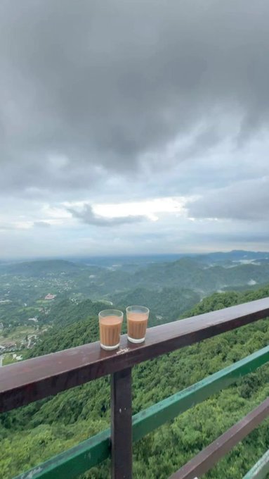 Two glasses of milk tea sit on a wooden railing overlooking a vast, cloudy green mountain landscape.