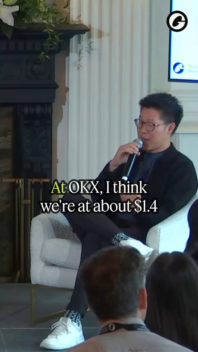 The image shows a man sitting in a chair, talking into a microphone during an event or presentation. The background features a TV screen displaying text related to the event, including phrases such as "At OKX", "I think we're at S14", and "Redeploy it in trading". There are also other people present in the scene, possibly listening to the man speaking into the microphone.