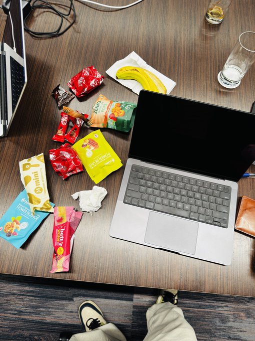 A laptop sits on a wooden desk surrounded by various snack wrappers, including "ZENGO," "omni," "PEARL GOURDE," and "Muesli" packets, alongside a banana on a napkin and water glasses, reflecting a casual workspace meal. The post's claim of a "wholesome lunch" is humorously contradicted by the abundance of packaged snacks and candy wrappers rather than a balanced meal.