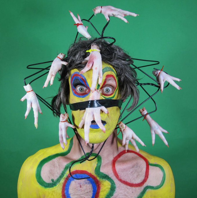 A person with yellow, green, red, and blue face and body paint wears a wire headpiece adorned with numerous rubber fingers, photographed against a green screen.