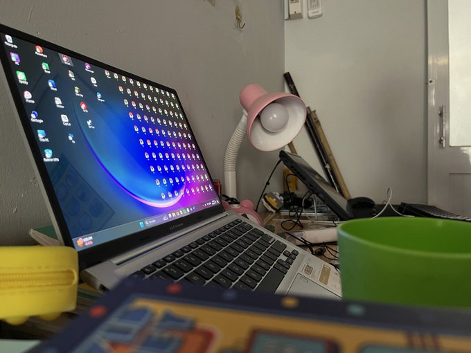 An angled view of a laptop with many desktop icons on screen, accompanied by a pink desk lamp, a green bowl, and a yellow banana on a cluttered desk.
