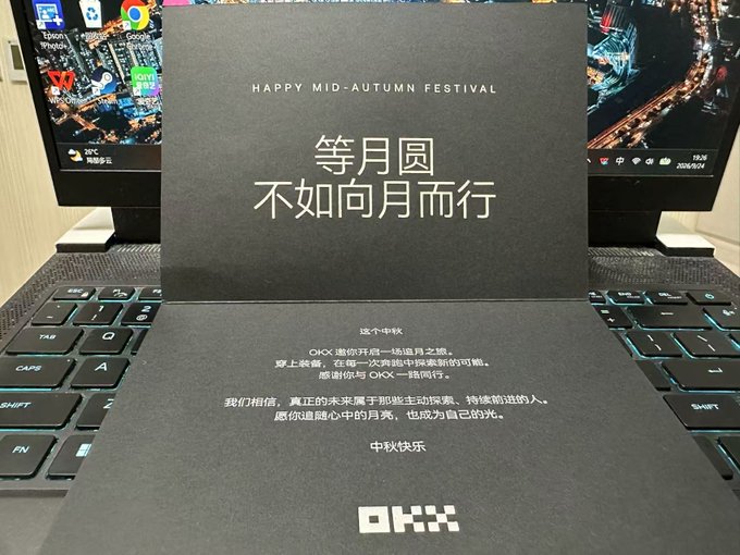 A dark OKX-branded Mid-Autumn Festival greeting card rests on a laptop keyboard, featuring the message "HAPPY MID-AUTUMN FESTIVAL" alongside the Chinese inscription "等月圆不如向月而行" (Don't wait for the moon to be full, walk toward the moon instead), with the OKX logo at the bottom—matching the recipient's praise for the thoughtful, well-designed holiday gift box from OKX.