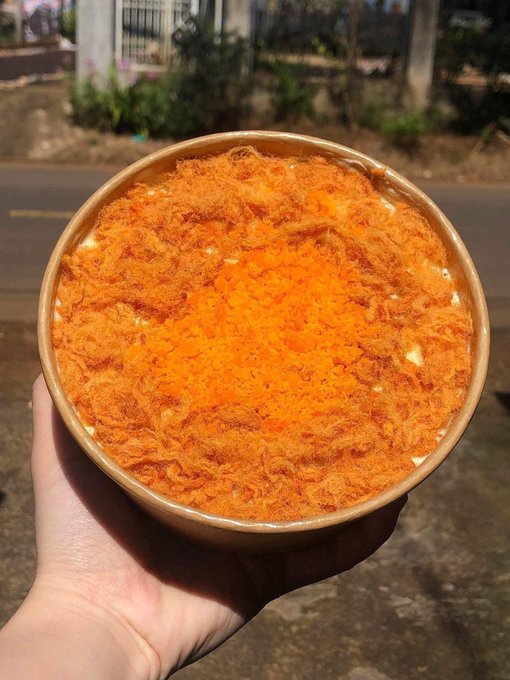 A hand holds up a round, brown paper container filled with a Vietnamese-style pizza topped with a generous, fluffy layer of shredded pork floss and orange cheese/sauce mixture, photographed outdoors in bright sunlight.

This complements the post, which casually shares a lazy weekend morning featuring this dish alongside coffee ("cf"), asking friends what they're having for breakfast ("ăn sáng món j").