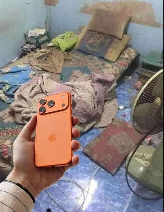 A hand holding an orange iPhone 17 Pro with a triple-camera design, photographed in a messy room with a worn mattress, blankets, and a fan.