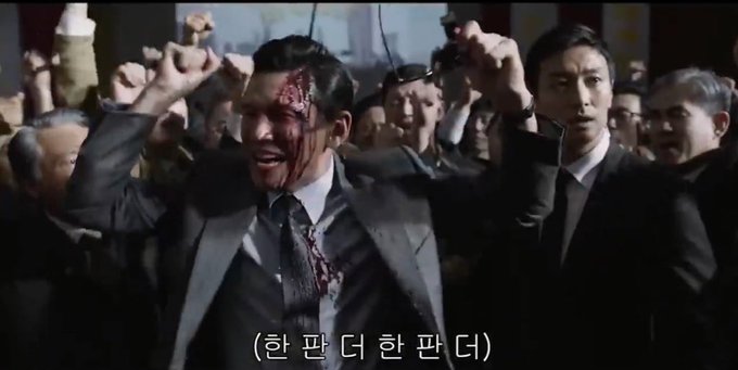 A scene from a film showing a bloody, injured man in a suit being held up by a crowd, with Korean subtitles reading '(한 판 더 한 판 더)' at the bottom.