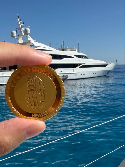A hand holding a gold commemorative coin with the text 'HEAD3' in front of a large luxury white yacht on blue ocean water.