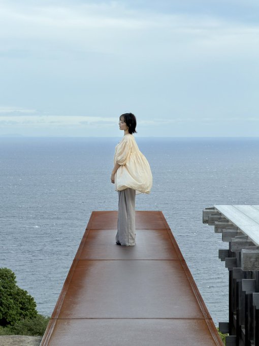 A woman stands contemplatively on a rust-colored walkway overlooking a vast ocean at Koyura Observatory, enjoying one of Tokyo's rare sunny days after weeks of overcast skies. Her pale yellow blouse catches the soft coastal breeze, while the steel-and-glass architecture frames the sweeping sea view, capturing the unexpected reprieve from the rainy weather.