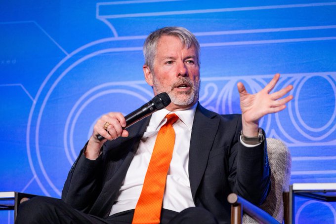 In the image, there is a man wearing a suit and an orange tie sitting in a chair with his hands outstretched. He appears to be talking into a microphone or addressing an audience at some event. The surrounding area seems to be designed for presentations or discussions. The man's hands are spread wide open, which might suggest that he is passionately explaining something related to the topic of Bitcoin. The content of his speech could be related to the price of Bitcoin or its potential impact on the market.