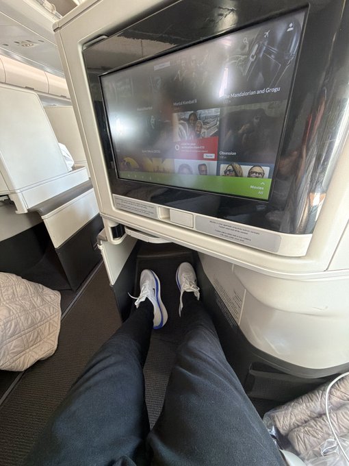 A first-person view from an airplane seat showing a passenger's legs stretched out in front of a seatback entertainment screen displaying a movie selection menu featuring 'Metal Combat 8' and 'Mandatorian and Gingu.'
