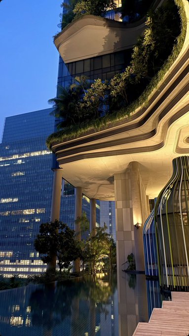 An evening cityscape featuring a modern, terraced building adorned with lush greenery and warm lighting, reflected in a serene pool alongside sleek glass skyscrapers. The luxurious, ambient setting evokes a stylish night out, perfectly complementing a fun evening spent with friends.