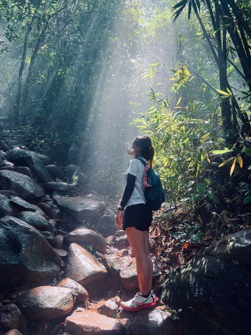 The image shows a woman standing on rocks near trees in a forest, wearing shorts and carrying a backpack. She appears to be looking up at something or someone above her. The photo has been captioned with a link to a post about going to the beach instead of the mountains, suggesting that she might have changed her plans.