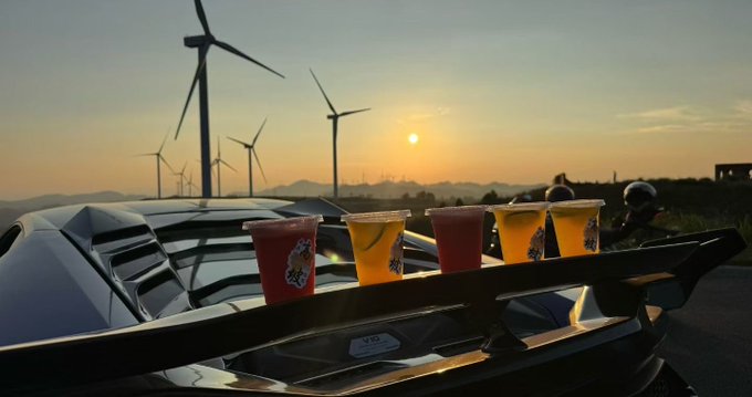 Four cups with colorful drinks sit on the hood of a dark car, with wind turbines silhouetted against a sunset sky in the background.