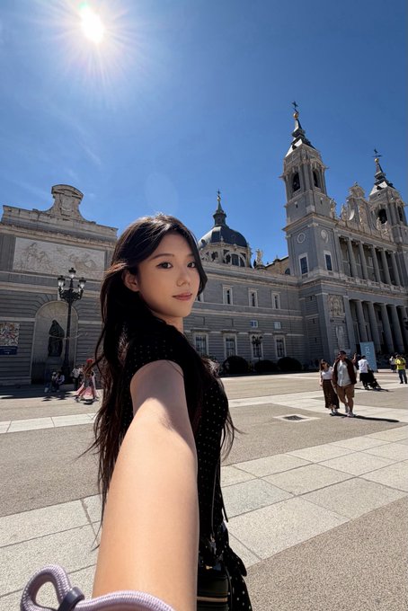 A selfie of a young Asian woman in a black dress taken in front of the Almudena Cathedral in Madrid, Spain, on a bright sunny day with tourists visible in the background plaza.