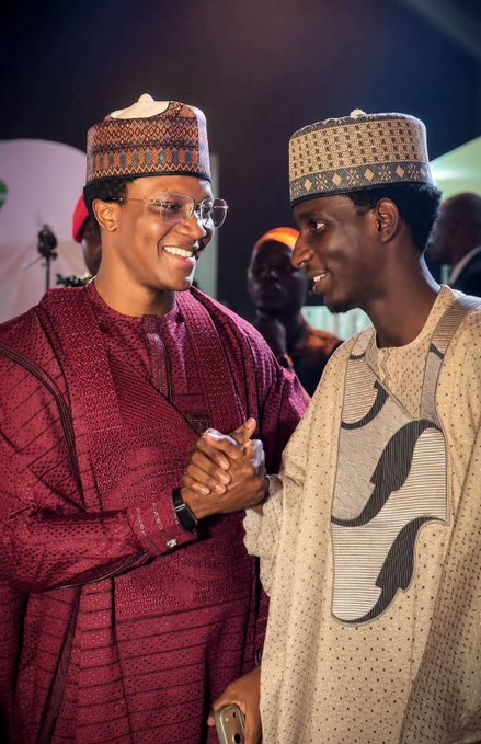 Two men in traditional Nigerian attire and caps smiling and clasping hands at an event.