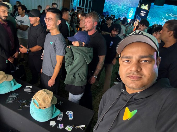 A selfie taken at a crowded gathering or conference, featuring a man in a black hoodie with a colorful logo and a black-and-white Adidas cap in the foreground, with other attendees and merchandise tables in the background.