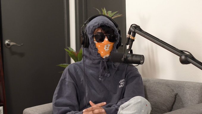 A person wearing a hoodie, sunglasses, an orange Bitcoin-themed face covering, and headphones sits on a couch in front of a professional podcast microphone. The image relates to the post by showing the subject, Frank DeGods, who transformed the poster's life after agreeing to a podcast collaboration in January 2022.