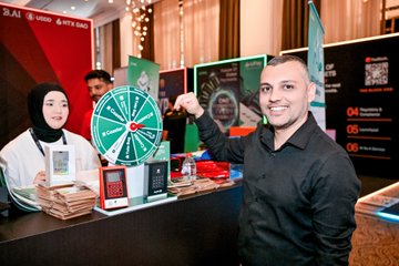 The image shows a man standing at a booth with a green spinning wheel, possibly participating in an event or game related to cryptocurrency. The booth has several items displayed, including a book titled "If you're wondering where the real energy is at #IBW2026", which could be about Web3 and its impact on the future of technology. There are also two people present at the booth, one standing next to it and another person in the background who appears to be a woman wearing a hijab. The scene suggests that this event or gathering might be related to cryptocurrency or Web3, attracting attendees interested in these topics.