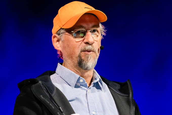 A middle-aged man with a gray beard, glasses, and an orange baseball cap speaks into a microphone against a deep blue backdrop, appearing to deliver a presentation. The image accompanies a quote from Adam Back predicting that Bitcoin could reach between $500,000 and $1,000,000 within the next 24 months.