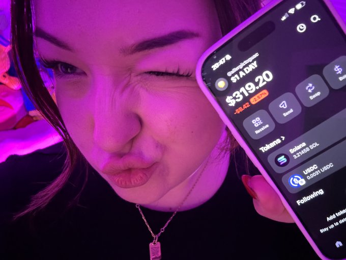 A close-up selfie of a young woman winking under pink/purple lighting, holding up a smartphone displaying a crypto portfolio screen showing $319.20 Solana balance with a -$8.42 (-2.57%) change and following tokens including USDC.