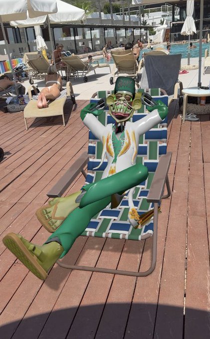 A 3D rendered zombie-like figure with a green cap, white jacket, and green pants reclining on a checkered deck chair by a pool area at a yacht club.