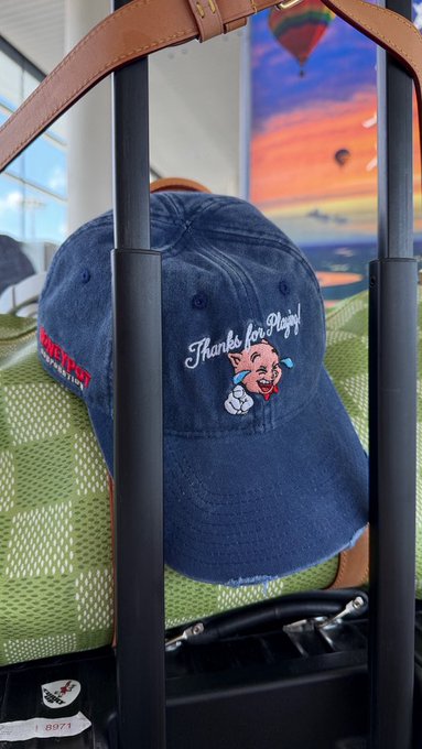 A blue denim cap featuring a 'Thanks for Playing!' embroidered patch with the Itchy & Scratchy cat and mouse, hanging on a luggage handle.