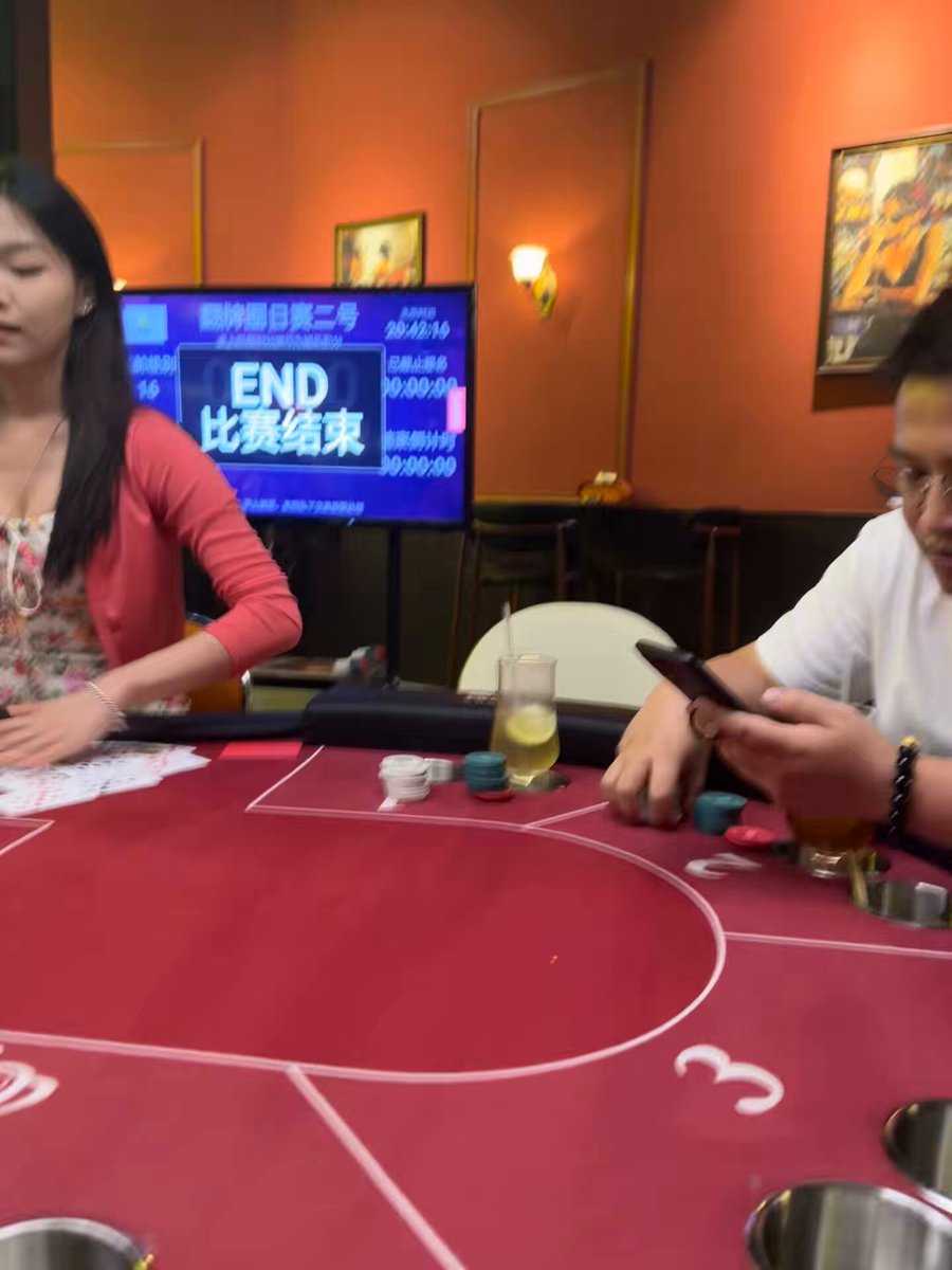 The image shows a woman sitting at a poker table, playing with a man who is also seated at the same table. The woman appears to be focused on the game while holding a cell phone in her hand. A TV screen can be seen behind them, displaying the text "END". There are two cups and a glass of water on the table as well. In the background, there is another person sitting at a different table with their own cell phone visible. The post suggests that the woman might have won $5000 in poker or has some other exciting news to share related to her winnings.