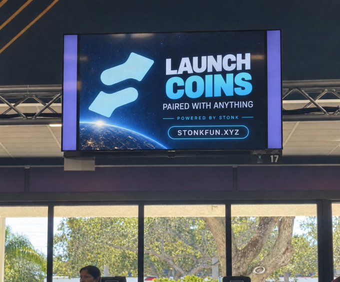 A digital billboard in an airport displaying an advertisement for 'LAUNCH COINS' powered by STONK with the website STONKFUN.XYZ.