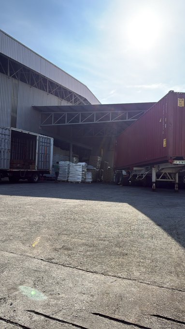 An outdoor industrial loading area at a warehouse with a red shipping container, an open container door revealing goods, and white sacks stacked inside under a curved metal roof.