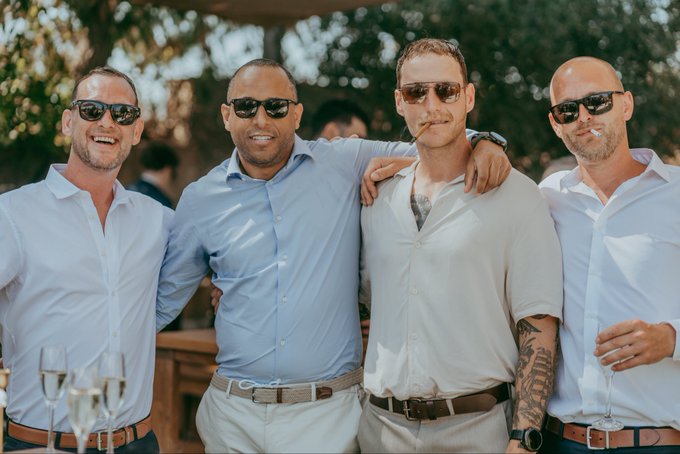 A group of four smiling men wearing sunglasses and light-colored button-up shirts pose together outdoors at what appears to be a social event, with one holding a glass of champagne.