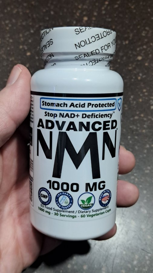 A hand holding a bottle of 'ADVANCED NMN 1000 MG' dietary supplement, labeled 'Stomach Acid Protected' and 'Stop NAD+ Deficiency', containing 60 vegetarian capsules.