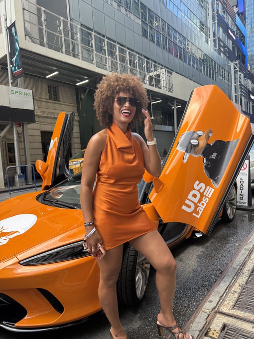 A woman in a short orange halter dress and sunglasses poses cheerfully next to an orange McLaren supercar with its doors open, branded with 'UDS Labs,' on a city street.