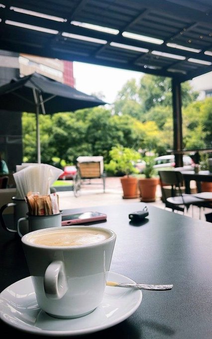 A cup of coffee on a saucer with a spoon, sitting on a dark table at an outdoor café under a pergola, with trees and umbrellas visible in the background.