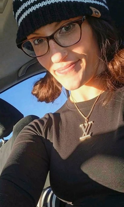 A woman wearing a navy and white striped beanie, glasses, a black top, and a gold Louis Vuitton (LV) necklace smiles softly in a selfie taken inside a vehicle.