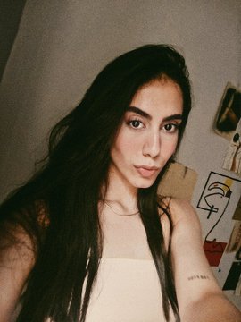 The image features a woman with long dark hair wearing a white tank top, posing for a photo. She has her arm around her shoulder and is looking directly at the camera. The background of the image includes a wall that appears to be gray or white in color. There are also some drawings on the wall behind the woman.