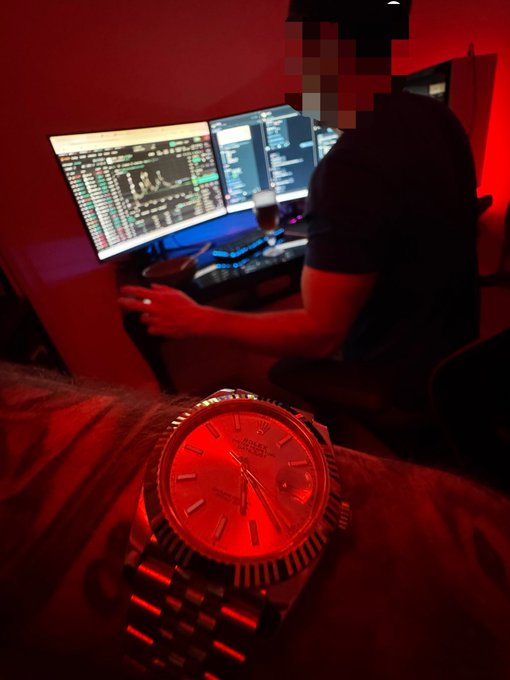 A red-tinted scene showing a luxury Rolex-style watch in the foreground with a person working on multiple trading monitors in the background.