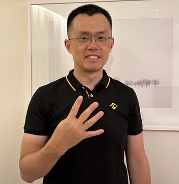 A man wearing glasses and a black polo shirt with a yellow logo smiles and gestures with his hand in front of a framed picture.