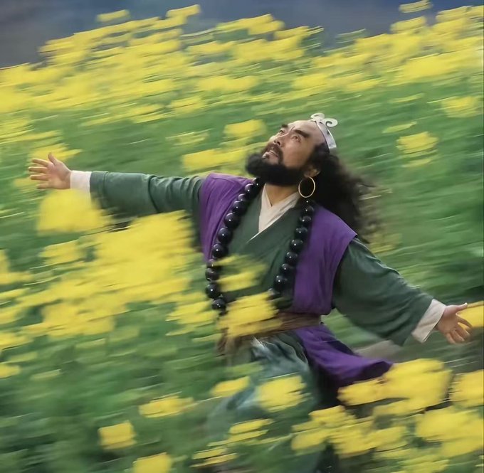 A man in traditional robes with a large beaded necklace and headpiece stands with arms outstretched among a field of yellow flowers, looking upward dramatically.
