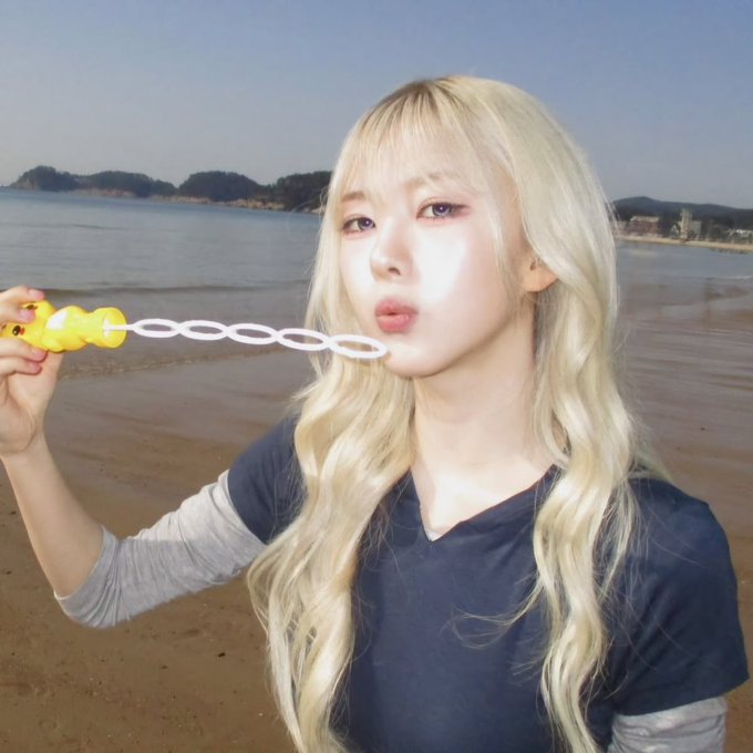 A blonde woman stands on a sandy beach blowing bubbles with a yellow wand, wearing a dark blue t-shirt over a grey long-sleeved shirt.