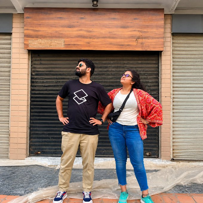 A photo of a man and a woman standing in front of a closed metal roller shutter; the man wears a black t-shirt with a white geometric logo and khaki pants, while the woman wears a red patterned cardigan, white top, blue jeans, and a black crossbody bag.