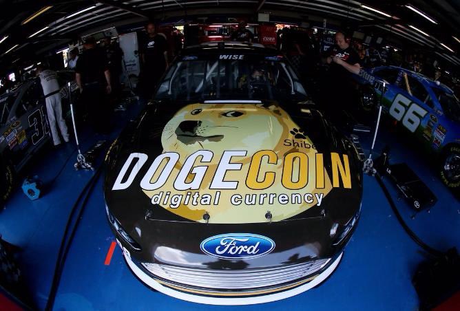 The image features a black race car with a yellow and white logo that reads "DOGE Coin" on its hood, indicating that it's a digital currency-themed vehicle. The car is parked inside a garage or warehouse, surrounded by other cars. There are several people in the background who appear to be either working on the vehicles or observing them.