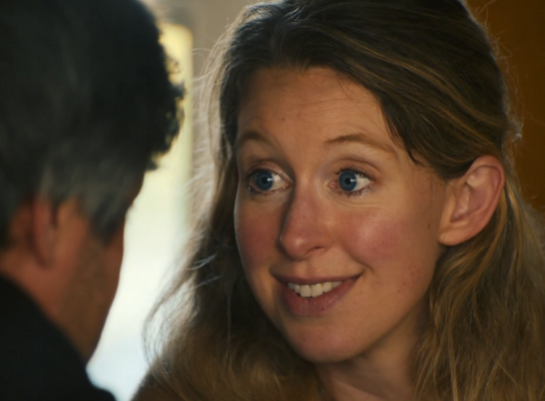 A close-up shows a woman with wide eyes and a forced, strained smile looking at a person whose back is turned to the camera, conveying an expression of nervous anticipation or feigned enthusiasm. The image perfectly captures the anxious, hopeful energy of someone reacting to a key opinion leader's callout on a low market cap cryptocurrency, where excitement is mixed with uncertainty.