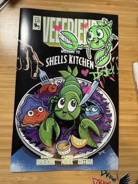 A photograph of a comic book titled 'VEGGIE FRIENDS: WELCOME TO SHELLS KITCHEN' featuring illustrated vegetable characters on a cover placed on a wooden surface.