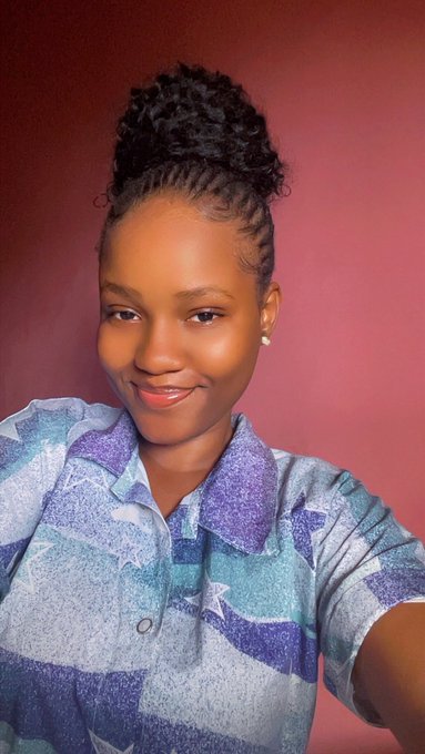 A young woman with a high curly bun takes a selfie against a pink wall, wearing a blue and purple patterned shirt and a pearl earring, smiling softly while participating in the "Day 63 of smiling" challenge for @ritualnet's Mainnet launch.