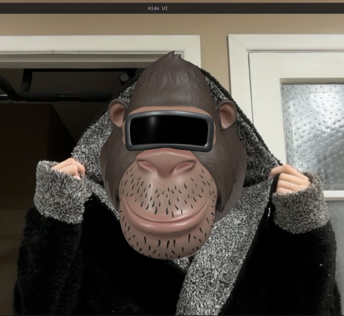 A person wearing a gray cardigan holds up their hood to reveal a Memoji-style gorilla face with a black VR headset overlaid on it, set in a dimly lit room.